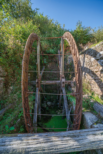 View of a large rusted iron water wheel set within the historic stone ruins and lush green vegetation of the Tregargus Valley Cornwall United Kingdom. View of a large rusted iron water wheel set within the historic stone ruins and lush green vegetation of the Tregargus Valley Cornwall United Kingdom.