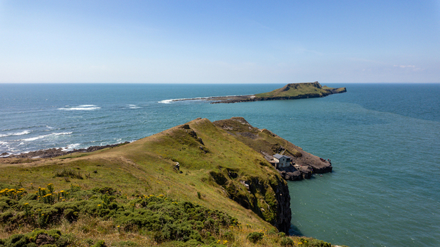 View of the grassy cliffs and the iconic Worm s Head headland stretching into the blue sea under a clear sky in Swansea Wales United Kingdom.