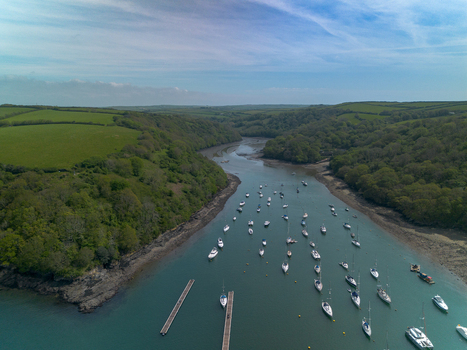 Aerial view of the Fowey River in Cornwall featuring numerous moored boats lush green hills and calm water under a bright blue sky Fowey Cornwall United Kingdom. Aerial view of the Fowey River in Cornwall featuring numerous moored boats lush green hills and calm water under a bright blue sky Fowey Cornwall United Kingdom.
