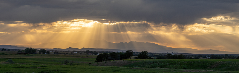 View of dramatic sun rays breaking through dark storm clouds over a mountain range and green fields at sunset United States. View of dramatic sun rays breaking through dark storm clouds over a mountain range and green fields at sunset United States.