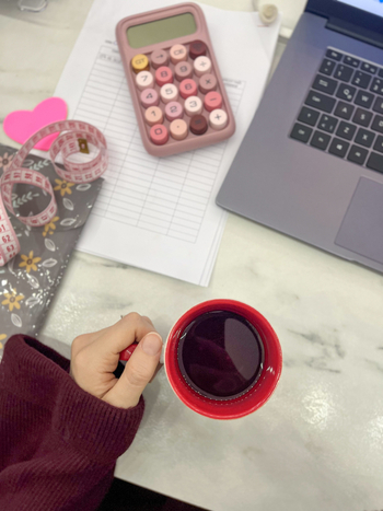 View of a person holding a red mug of tea at a desk with a laptop pink calculator measuring tape and documents on a marble surface in Beylikduzu Istanbul Turkiye. View of a person holding a red mug of tea at a desk with a laptop pink calculator measuring tape and documents on a marble surface in Beylikduzu Istanbul Turkiye.