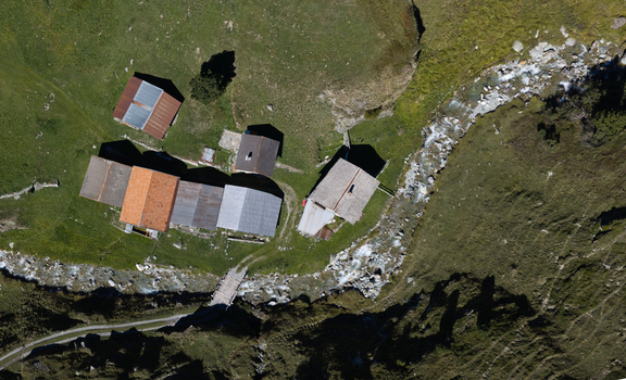 Aerial view of a cluster of traditional alpine farmhouses with orange and grey roofs situated beside a small mountain stream and a stone bridge in the green meadows of Bivio Grisons Switzerland. Aerial view of a cluster of traditional alpine farmhouses with orange and grey roofs situated beside a small mountain stream and a stone bridge in the green meadows of Bivio Grisons Switzerland.