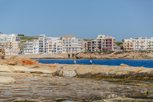 View of sun drenched rocks blue water and buildings under a clear sky create a vibrant coastal scene Marsaskala Malta. View of sun drenched rocks blue water and buildings under a clear sky create a vibrant coastal scene Marsaskala Malta.