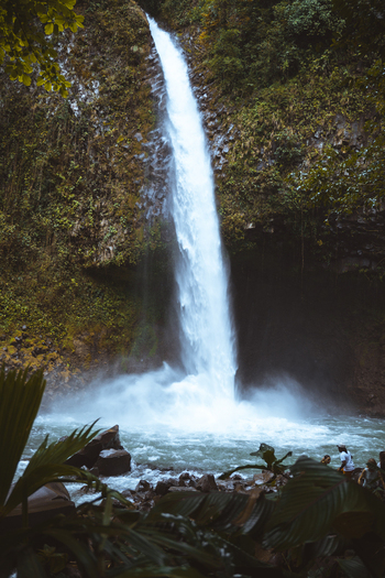 View of a powerful waterfall cascading down a lush moss covered cliff into a rocky pool with people standing nearby in Costa Rica.