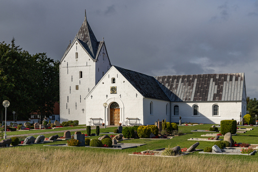 View of Romo Church featuring its white facade and pointed tower surrounded by a cemetery with gravestones under a cloudy sky in Romo Denmark. View of Romo Church featuring its white facade and pointed tower surrounded by a cemetery with gravestones under a cloudy sky in Romo Denmark.