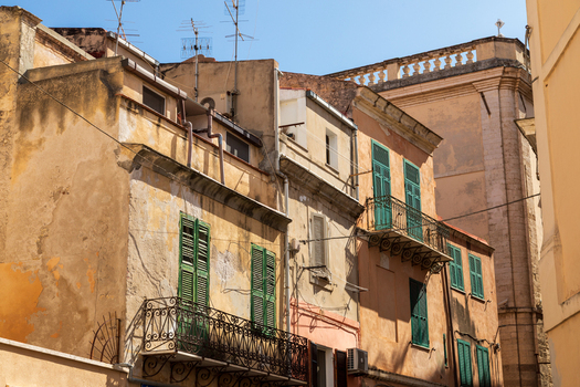 View of historic residential buildings with weathered facades green shutters and ornate wrought iron balconies in Sardinia Italy.