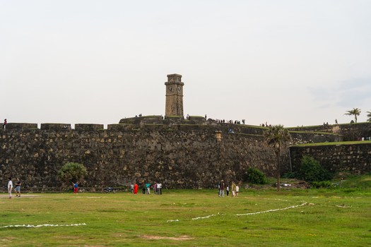 View of the historic Galle Fort Clock Tower rising above massive stone ramparts with people walking on the grass and walls Galle Southern Province Sri Lanka.