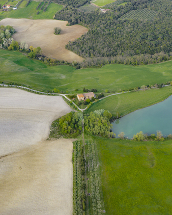Aerial view of a traditional farmhouse nestled among rolling green hills cultivated fields and a small turquoise lake Pomarance Tuscany Italy.