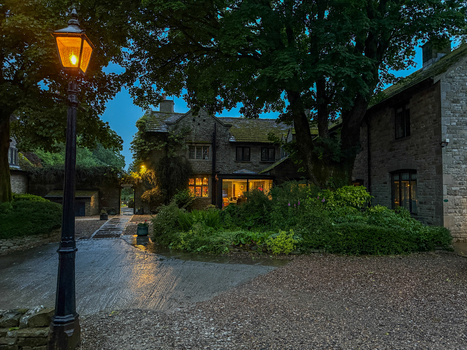 View of a traditional stone cottage and a lit vintage street lamp at twilight with lush green trees and warm glowing windows in Hawes England United Kingdom. View of a traditional stone cottage and a lit vintage street lamp at twilight with lush green trees and warm glowing windows in Hawes England United Kingdom.