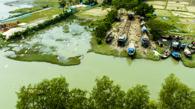 Aerial view of traditional wooden boats on a slipway along the riverbank with lush green vegetation and white birds flying over the water in Patharghata Barisal Division Bangladesh. Aerial view of traditional wooden boats on a slipway along the riverbank with lush green vegetation and white birds flying over the water in Patharghata Barisal Division Bangladesh.