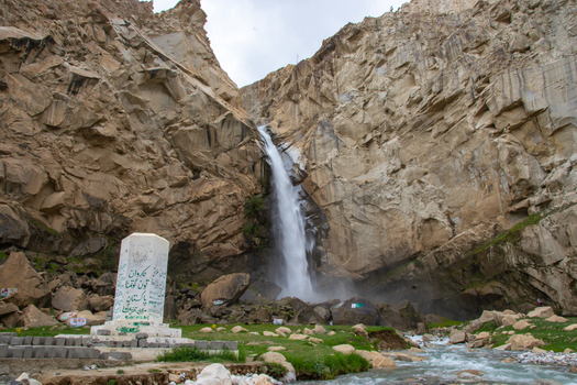 View of Khamosh Waterfall cascading down steep rocky cliffs with a white monument in the foreground in Skardu Gilgit Pakistan.