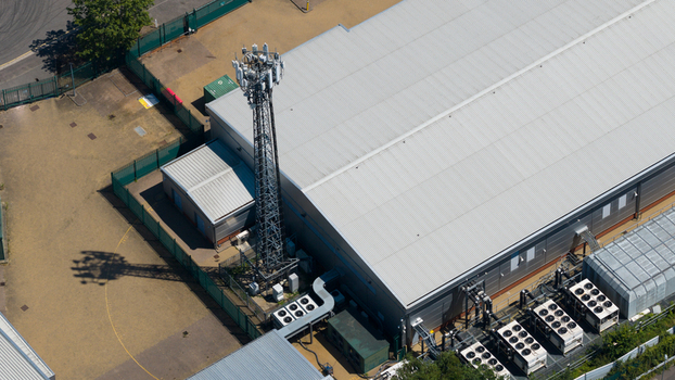 Aerial view of a telecommunications tower situated next to a large industrial facility featuring rooftop HVAC units and fenced perimeter in Hemel Hempstead England United Kingdom.