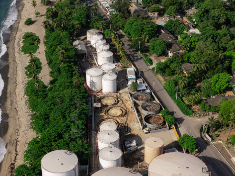 Aerial view of industrial storage tanks situated along a coastline with lush tropical trees and a sandy beach in Bajos de Haina San Cristobal Provinc Dominican Republic.