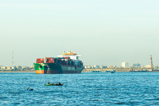 View of a large container ship sailing near the coast with small fishing boats carrying local fishermen in the foreground and the city skyline in Karachi Sindh Pakistan.