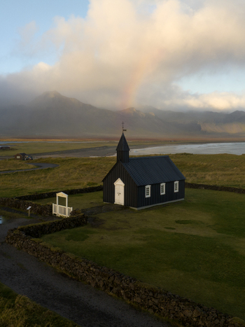 Aerial view of the iconic black church standing stark against the green landscape with a faint rainbow B&uacute;&eth;ir Sn&aelig;fellsb&aelig;r Iceland.
