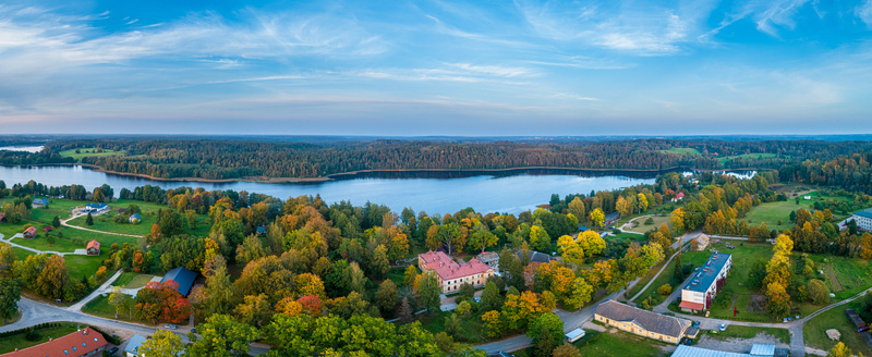 Aerial view of a serene lake surrounded by dense autumn forests and scattered buildings under a clear blue sky in Latvia. Aerial view of a serene lake surrounded by dense autumn forests and scattered buildings under a clear blue sky in Latvia.