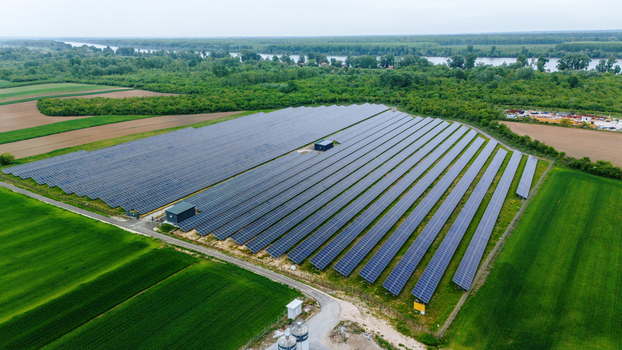Aerial view of a large solar panel array surrounded by lush green agricultural fields and dense forest near a river in Sabac Vojvodina Serbia.
