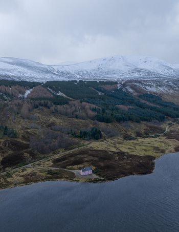 Aerial view of The Pink House on the shore of Loch Glass with snow capped mountains and evergreen forests under a cloudy sky in Dingwall Scotland United Kingdom. Aerial view of The Pink House on the shore of Loch Glass with snow capped mountains and evergreen forests under a cloudy sky in Dingwall Scotland United Kingdom.
