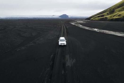 Aerial view of a lone white car driving through the stark black volcanic sands towards the distant icy glacial Maelifell Iceland. Aerial view of a lone white car driving through the stark black volcanic sands towards the distant icy glacial Maelifell Iceland.