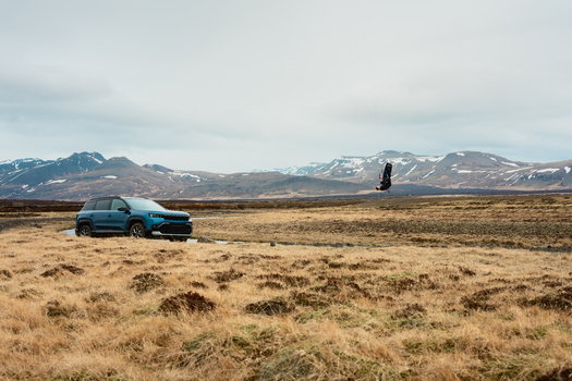 View of a blue SUV parked on a dirt road in a vast dry grassy field with a person kiteboarding in the air against a backdrop of snow capped mountains in Borgarbyggd Iceland. View of a blue SUV parked on a dirt road in a vast dry grassy field with a person kiteboarding in the air against a backdrop of snow capped mountains in Borgarbyggd Iceland.