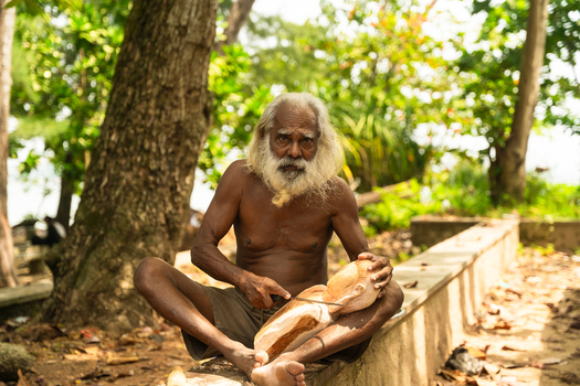 Sri Lanka 12 February 2026 View of an elderly man with a long white beard carving a wooden sculpture while sitting outdoors under the shade of trees.