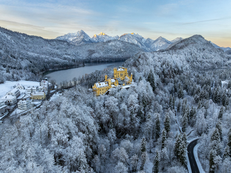 Aerial view of Hohenschwangau Castle nestled among snow covered trees with a frozen lake and mountains in the distance Schwangau Bayern Germany.