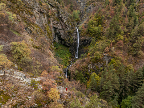 Aerial view of Ovcharenski Waterfall Goritsa surrounded by autumn foliage in a rocky gorge with a person in a red jacket standing on a trail in Ovchartsi Kyustendil Province Bulgaria. Aerial view of Ovcharenski Waterfall Goritsa surrounded by autumn foliage in a rocky gorge with a person in a red jacket standing on a trail in Ovchartsi Kyustendil Province Bulgaria.