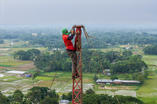 Trishal Upazila Bangladesh 13 May 2026 Aerial view of a worker climbing a red metal tower using ropes and pulleys over a lush green rural landscape with agricultural fields. Trishal Upazila Bangladesh 13 May 2026 Aerial view of a worker climbing a red metal tower using ropes and pulleys over a lush green rural landscape with agricultural fields.