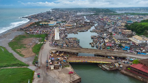 Aerial view of the harbor featuring wooden fishing boats docked along the river a bridge and the surrounding coastal town with dense rooftops in Elmina Central Region Ghana. Aerial view of the harbor featuring wooden fishing boats docked along the river a bridge and the surrounding coastal town with dense rooftops in Elmina Central Region Ghana.