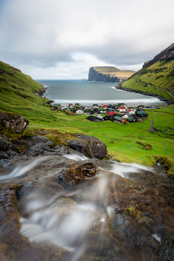 View of the waterfall at Tjornuvik cascading over mossy rocks with a village nestled in a green valley by the ocean in the Faroe Islands.