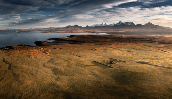 Aerial view of the Budir Black Church in a vast grassy field near the coast with snow capped mountains under a dramatic sky in Iceland.