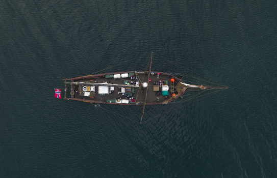 Aerial view of a traditional wooden sailing ship with a Norwegian flag and people on deck navigating the dark waters of Hardangerfjord in Blurnes Vestland Norway. Aerial view of a traditional wooden sailing ship with a Norwegian flag and people on deck navigating the dark waters of Hardangerfjord in Blurnes Vestland Norway.