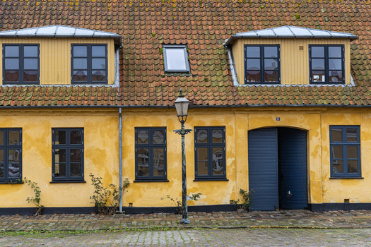 View of a traditional yellow walled building with a red tiled roof dark window frames and a vintage street lamp in front of a cobblestone street in Praesto Seeland Denmark. View of a traditional yellow walled building with a red tiled roof dark window frames and a vintage street lamp in front of a cobblestone street in Praesto Seeland Denmark.