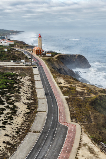 Aerial view of Farol Penedo da Saudade lighthouse standing on rugged cliffs beside a coastal road and crashing Atlantic waves in Marinha Grande Leiria Portugal. Aerial view of Farol Penedo da Saudade lighthouse standing on rugged cliffs beside a coastal road and crashing Atlantic waves in Marinha Grande Leiria Portugal.