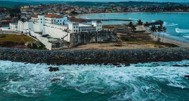 Aerial view of Elmina Castle a historic white fortress situated on the coast with crashing waves palm trees and the town in the background Ghana. Aerial view of Elmina Castle a historic white fortress situated on the coast with crashing waves palm trees and the town in the background Ghana.
