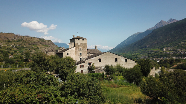Aerial view of a medieval castle surrounded by lush green trees and vineyards with majestic mountains in the background under a clear blue sky Aosta Valley Aosta Valley Italy. Aerial view of a medieval castle surrounded by lush green trees and vineyards with majestic mountains in the background under a clear blue sky Aosta Valley Aosta Valley Italy.