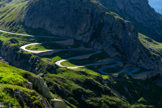View of the winding serpentine road of the Sankt Gotthard Pass carved into steep rocky mountain slopes and lush green alpine meadows in Airolo Ticino Switzerland.