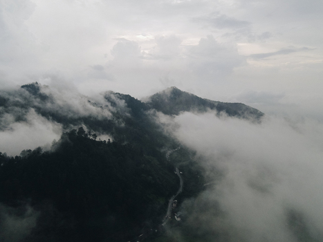 Aerial view of misty mountains and lush green slopes partially covered by thick white clouds with a winding road below under an overcast sky in Tawangmangu Central Java Indonesia.