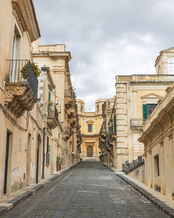 View of a narrow cobblestone street lined with ornate Baroque buildings and balconies leading to a historic church under an overcast sky in Noto Sicily Italy. View of a narrow cobblestone street lined with ornate Baroque buildings and balconies leading to a historic church under an overcast sky in Noto Sicily Italy.