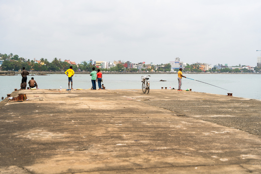 Sri Lanka 07 January 2026 View of people standing on a concrete pier with a man fishing and a coastal town skyline in the background.