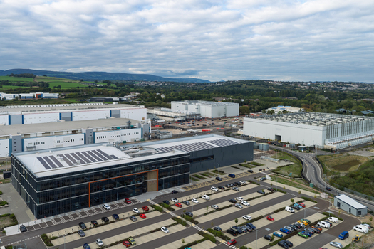 Aerial view of a large data center complex featuring modern industrial buildings with solar panels on the roofs surrounded by parking lots and green landscape in the United Kingdom.
