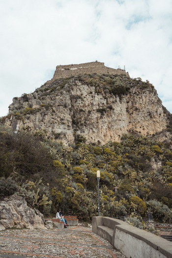 View of a majestic fortress perched atop a rugged rocky mountain its weathered stone contrasting with the vibrant green foliage below Taormina Sicily Italy.