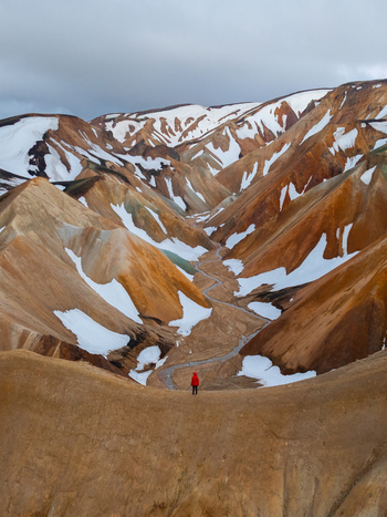 Aerial view of Landmannalaugar featuring colorful rhyolite mountains with patches of snow and a winding stream with a person in a red jacket standing on a ridge in Rangarthing ytra Iceland. Aerial view of Landmannalaugar featuring colorful rhyolite mountains with patches of snow and a winding stream with a person in a red jacket standing on a ridge in Rangarthing ytra Iceland.