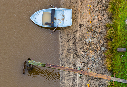 Aerial view of a small boat moored near a wooden pier on a muddy riverbank in Latvia.