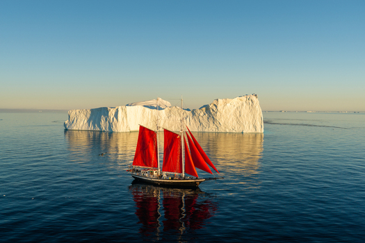 Aerial view of a sailboat with striking red sails navigating calm blue waters in front of a massive white iceberg under a clear sky in Greenland. Aerial view of a sailboat with striking red sails navigating calm blue waters in front of a massive white iceberg under a clear sky in Greenland.