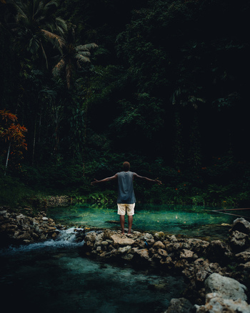 View of a lone figure stands poised on the rocky edge of a turquoise pool arms outstretched embraced by the emerald depths of the surrounding forest Lata Solomon Islands.