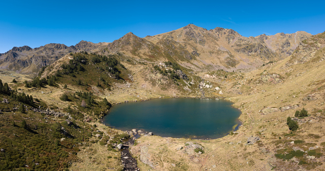 Aerial view of Tristaina Lakes surrounded by rugged mountain peaks and grassy slopes under a clear blue sky in Ordino Andorra.