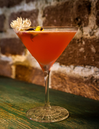 View of a pink cocktail with edible flowers in a martini glass on a green wooden counter against a brick wall at Ruba s Bakery Bistro San Jose del Cabo Baja California Sur Mexico.