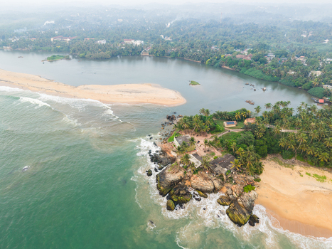 Aerial view of the coastline where a river estuary meets the turquoise ocean waves near a rocky headland with palm trees and sandy beaches in Galle Southern Province Sri Lanka.