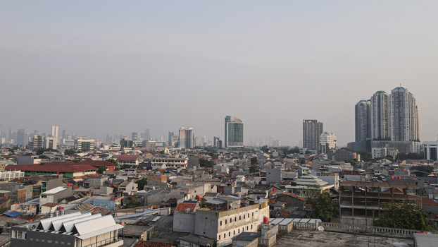 Aerial view of the dense urban landscape featuring a mix of low rise residential buildings and modern high rise skyscrapers under a hazy sky in West Jakarta Jakarta Indonesia.